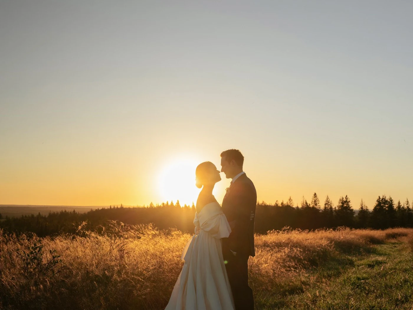 Bride and groom standing close in silhouette against a low sun in an open field.