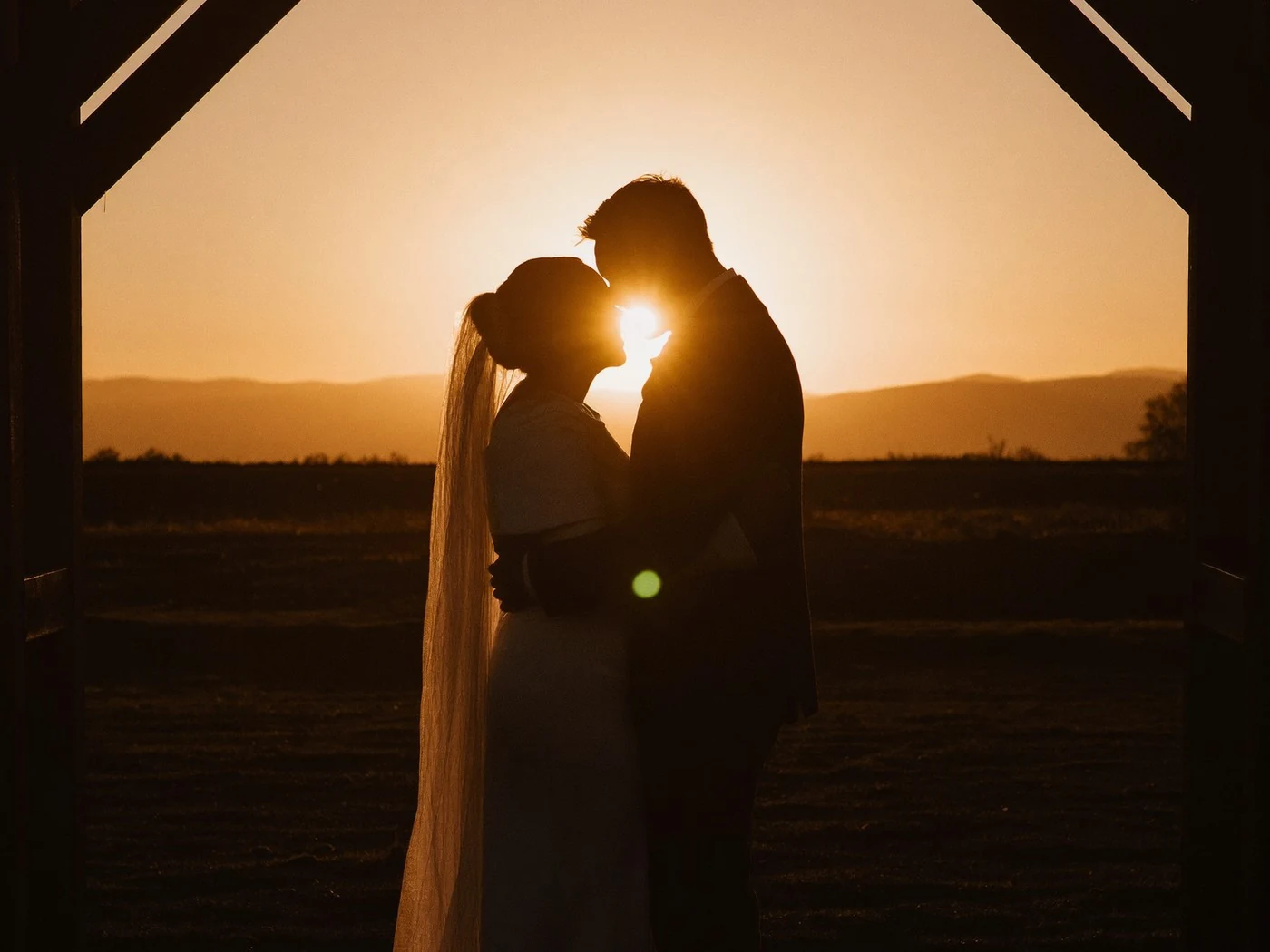 Couple silhouetted under a timber arch about to kiss, setting sun glowing between them.