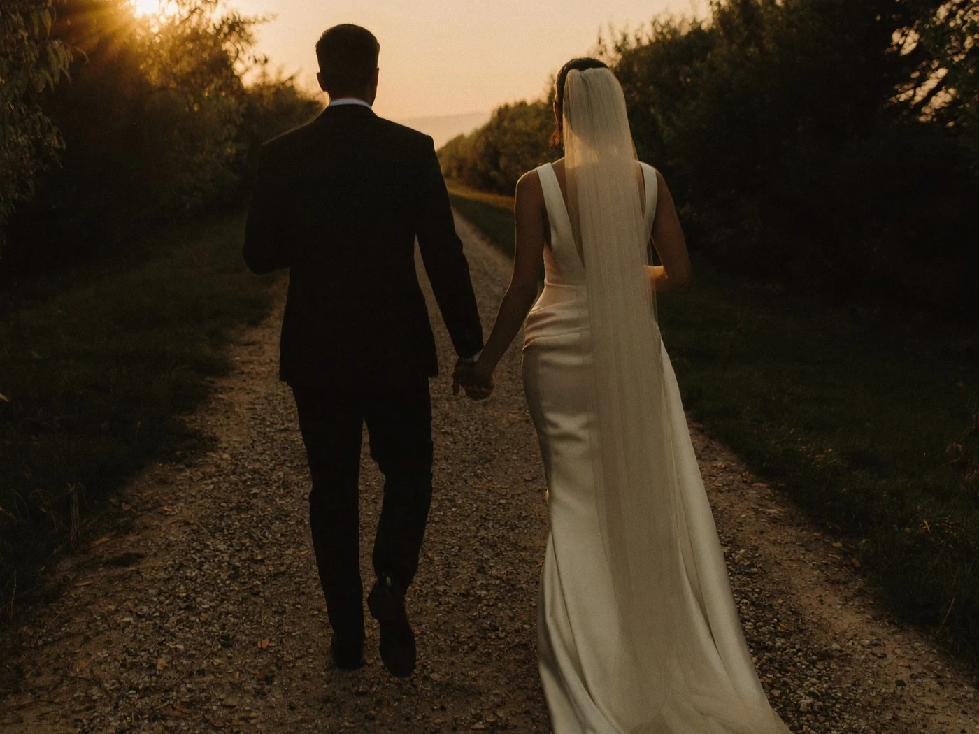 Bride's long veil and satin train sweep a narrow gravel track between dark hedgerows.