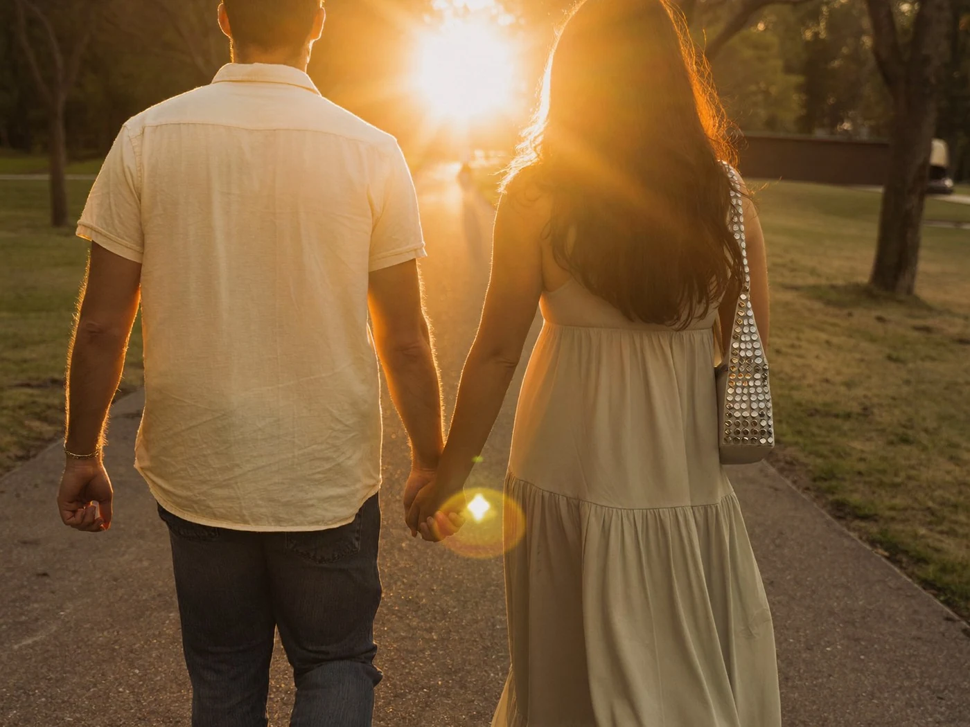 Couple in jeans and a sundress walking a park path into a sun starburst.