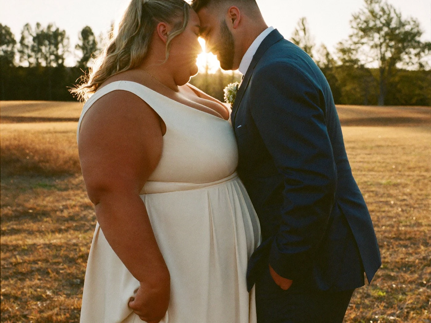 Plus size bride and groom forehead to forehead in a golden field, backlit by a low sun.