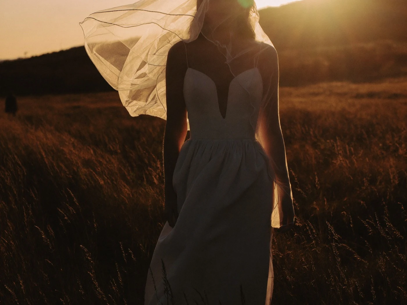 Bride standing in tall grass at sunset, her veil lifted sideways by the wind, backlit.