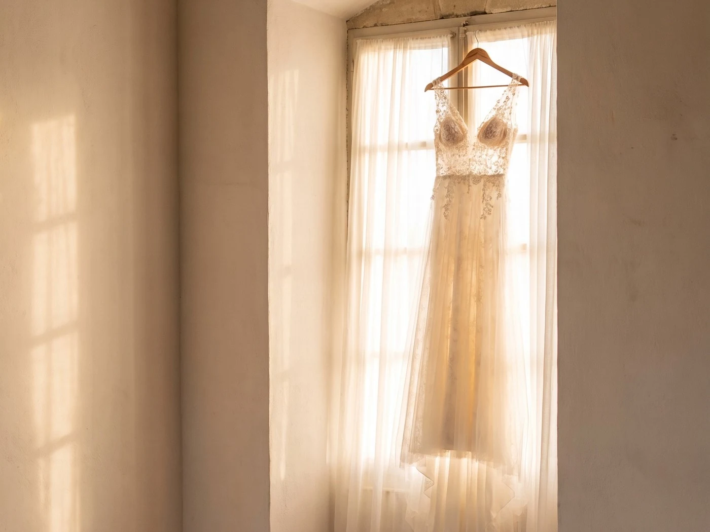 Ivory lace wedding gown hanging in a tall arched window, sheer curtains and warm light.