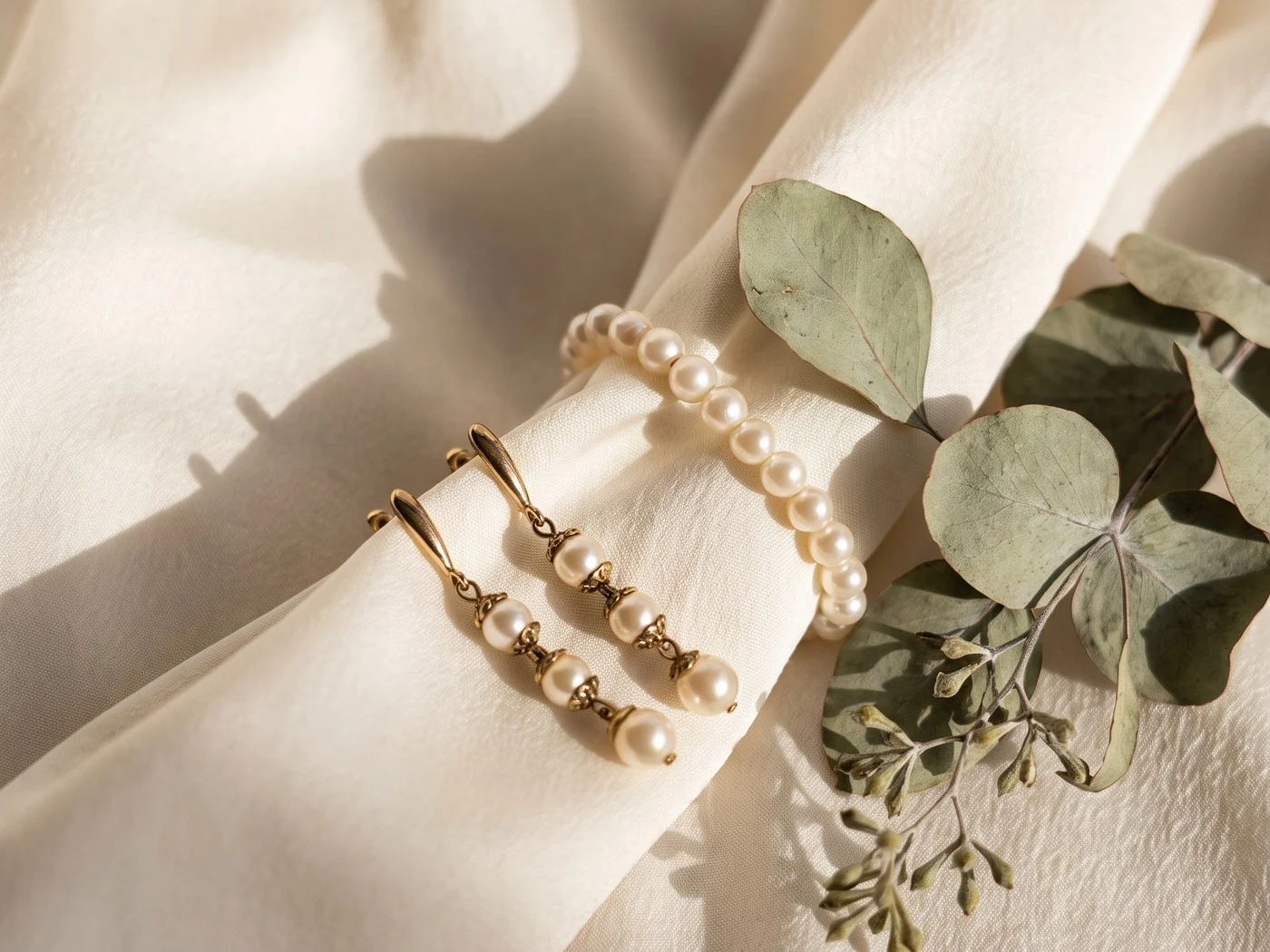 Pearl drop earrings and a pearl bracelet resting on cream silk beside a eucalyptus sprig.