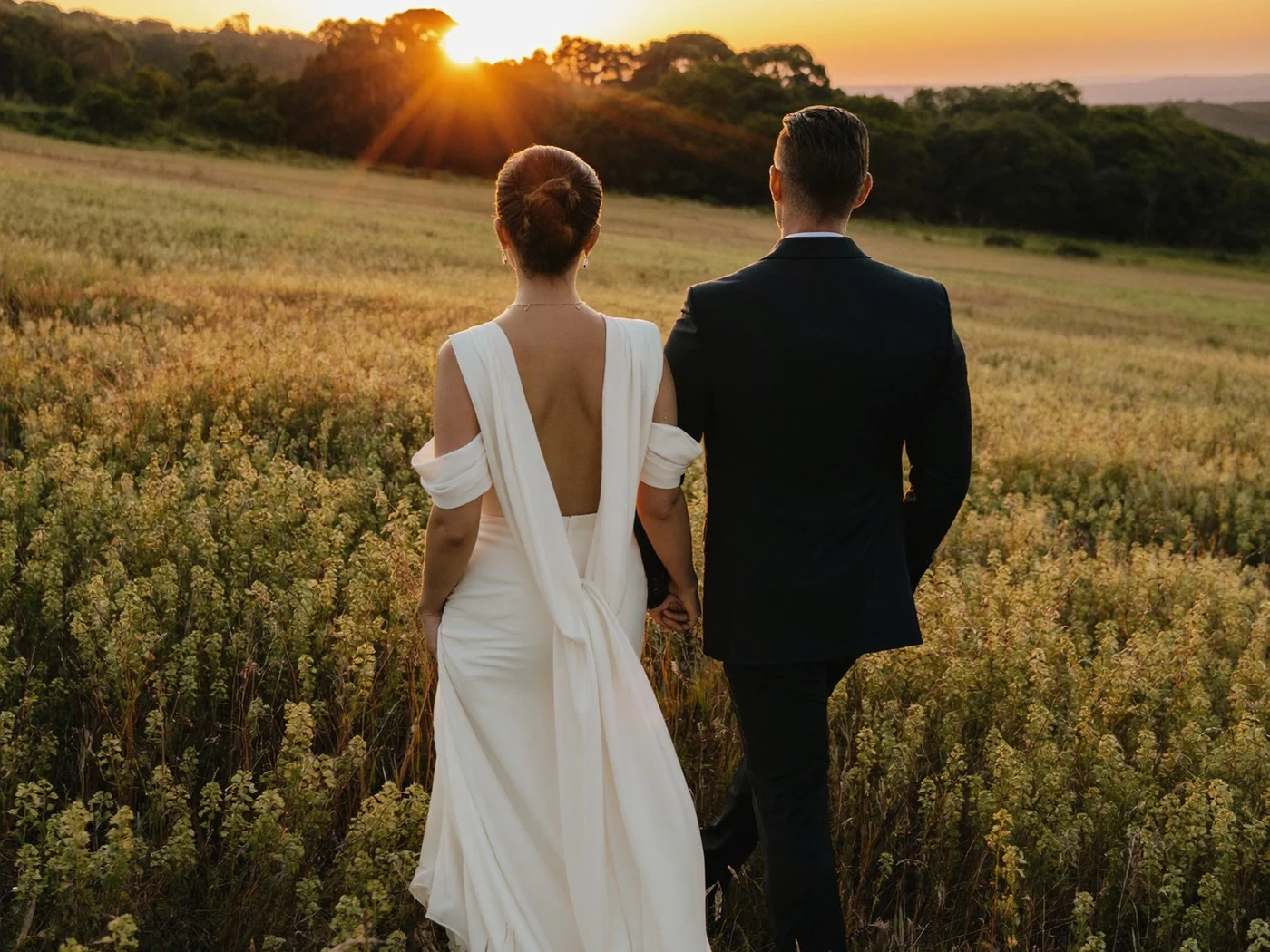 Bride and groom seen from behind walking through a golden wildflower meadow at sunset.
