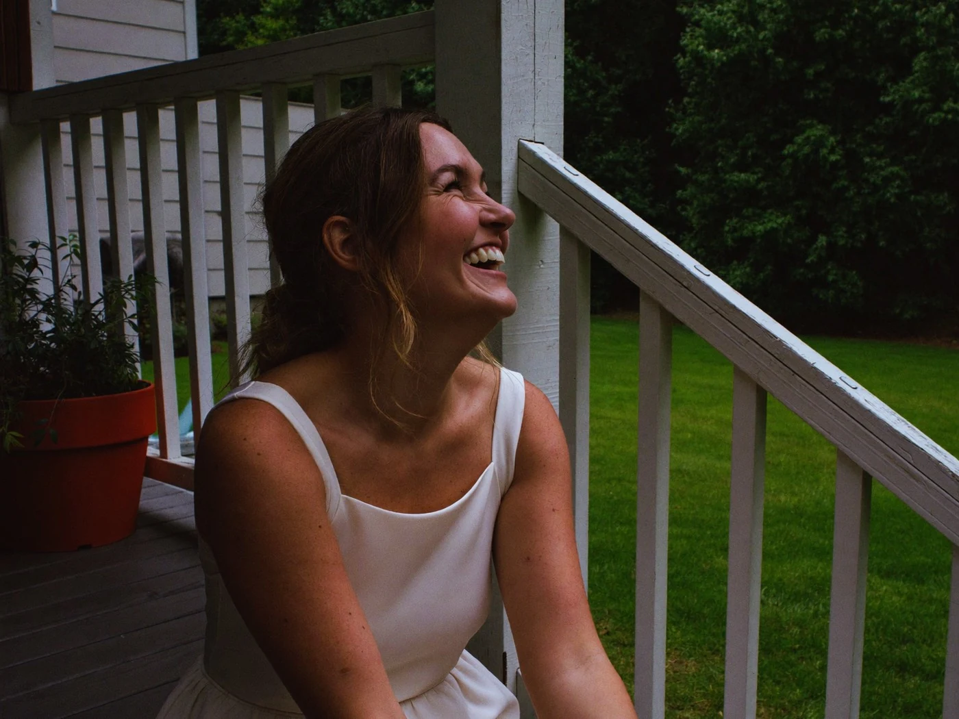 Bride in a simple ivory dress laughing on wooden porch steps, white railing and green lawn behind.