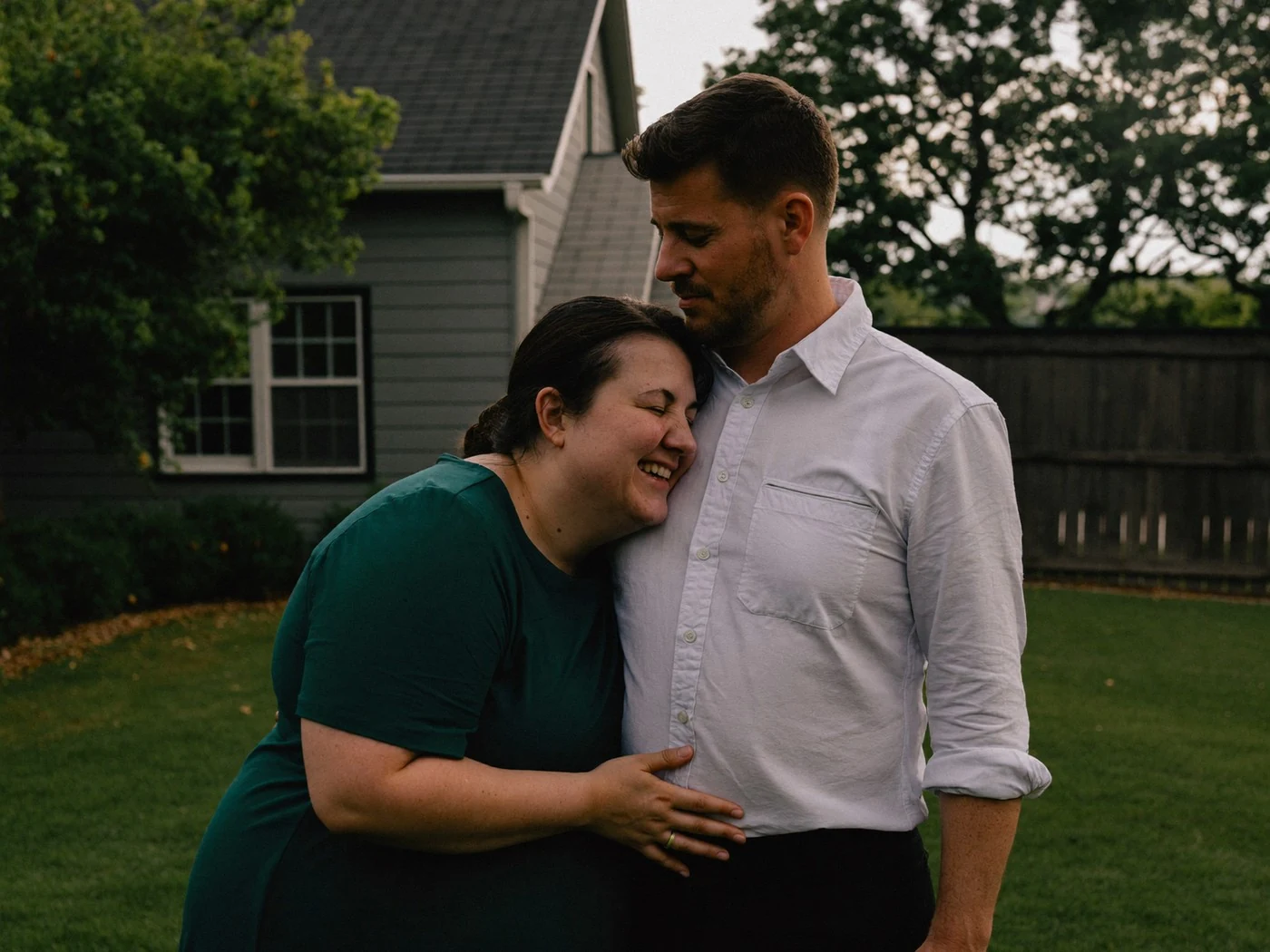 Couple standing close on a suburban back lawn, her head resting on his shoulder, house and fence behind.