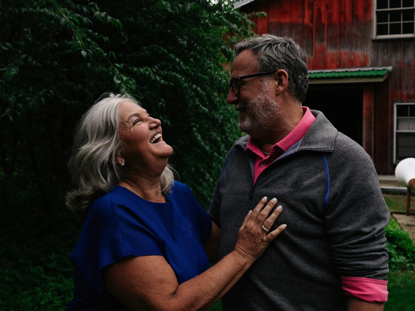 Couple in their fifties laughing together in a green backyard beside a weathered red barn.