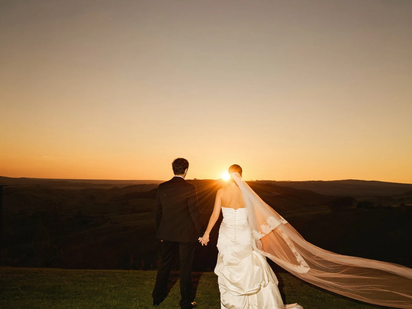 Couple stand still above a wide valley, her long veil blown horizontally across frame.