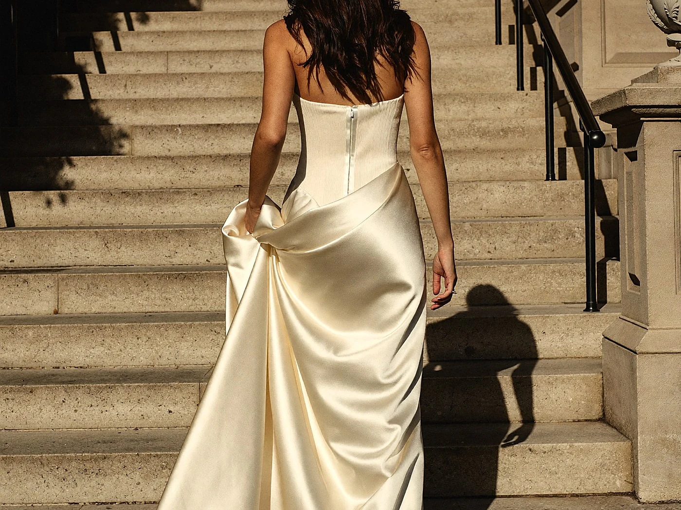 Bride from behind climbing wide stone steps, ivory satin gown and train sweeping behind her.