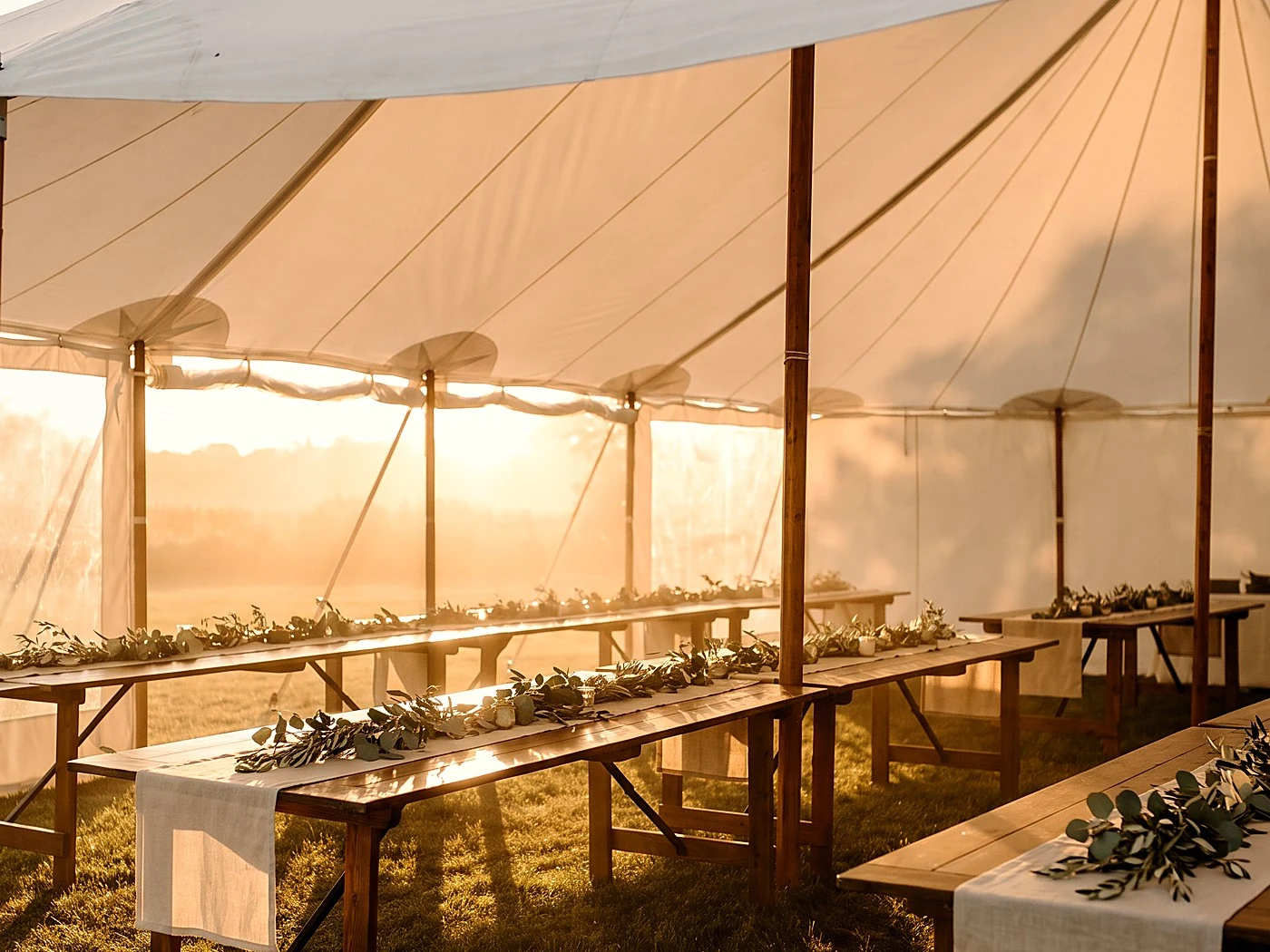 Wooden farm tables with linen runners and greenery inside a white tent at golden sunset.