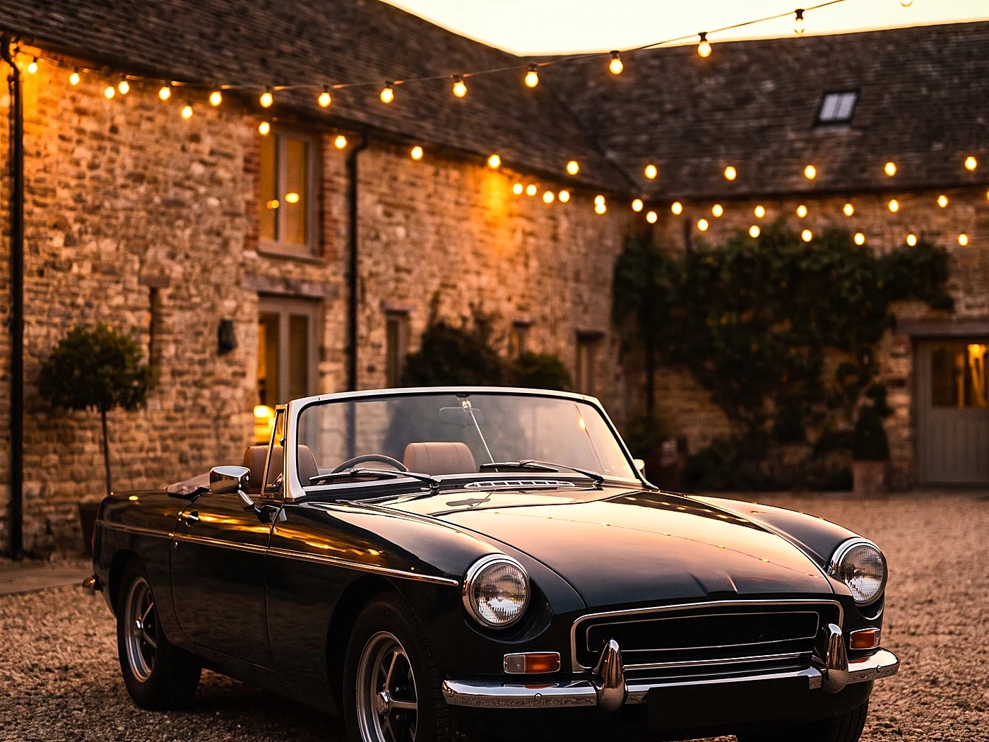 Dark green vintage convertible parked on gravel below string lights, stone courtyard walls behind.
