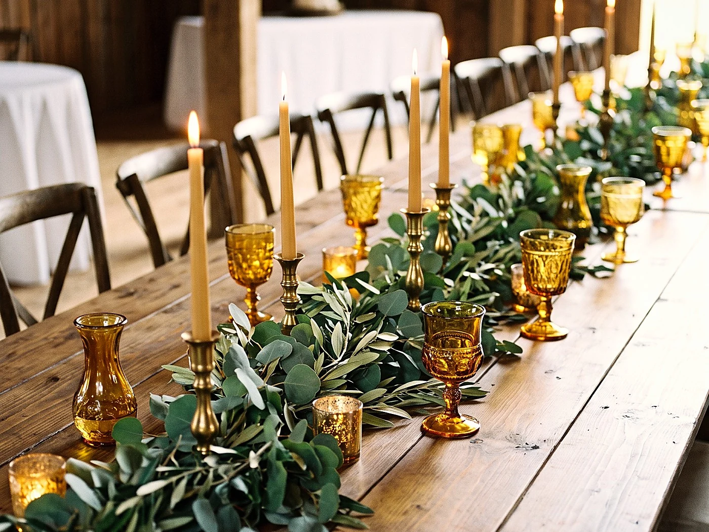 A long farmhouse table with greenery runner, brass candlesticks and amber glass, shot low along its length.