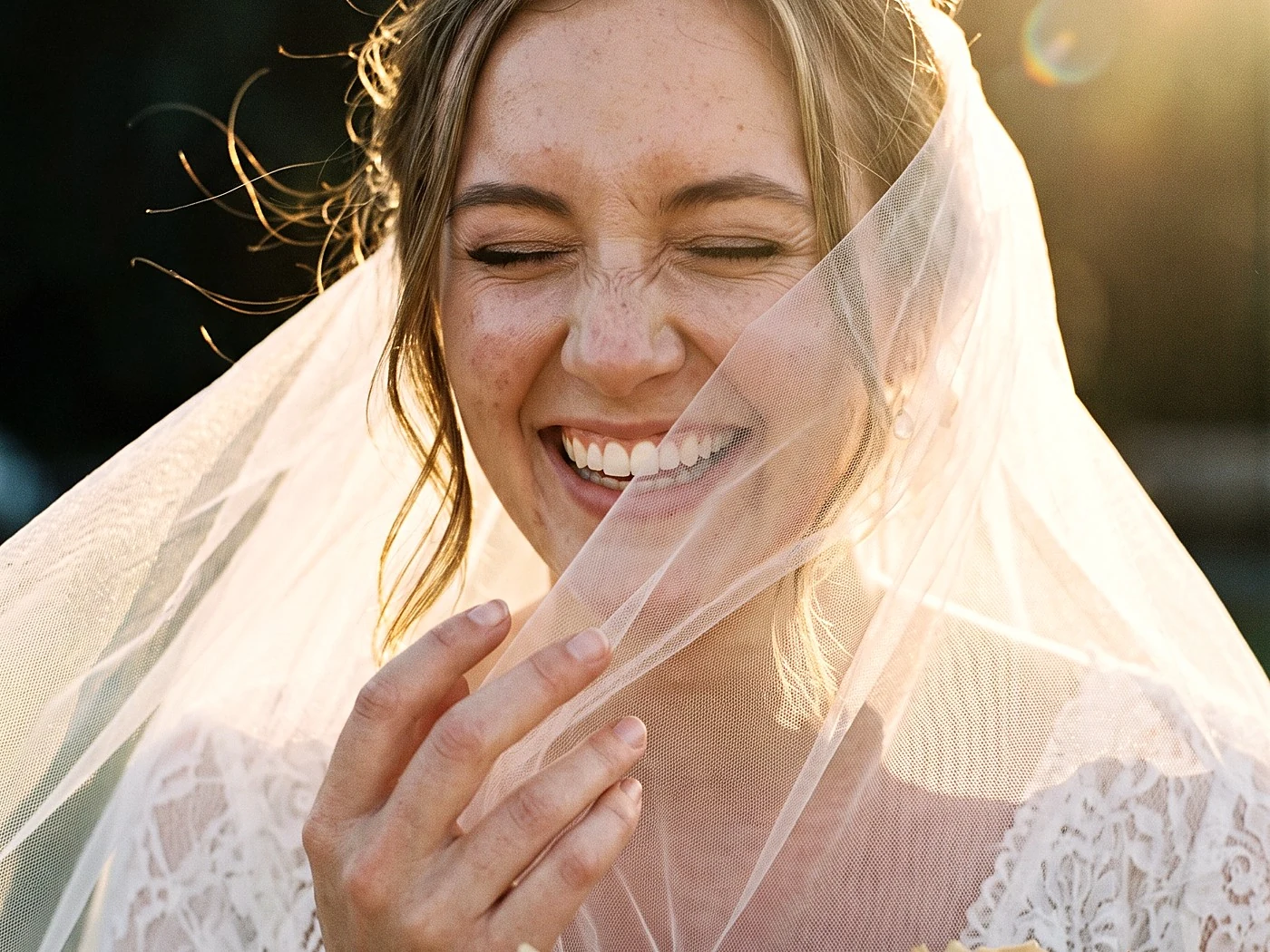 Close portrait of a bride in her late twenties laughing with eyes closed, veil catching wind, warm rim light.