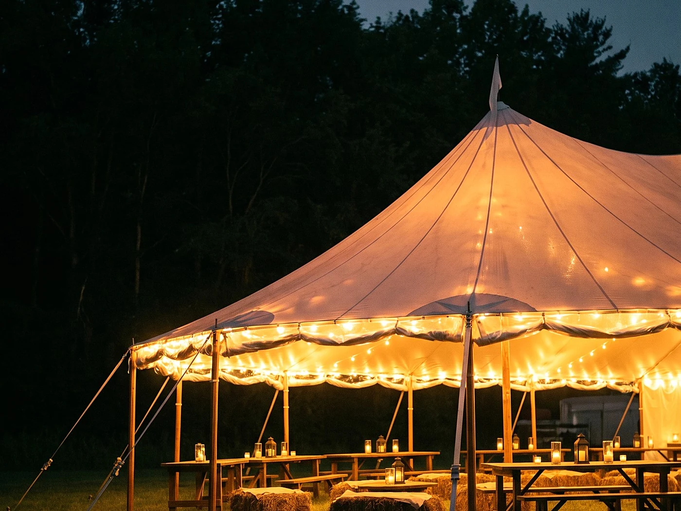 A sailcloth tent glowing from within at night, string lights, dark treeline behind.