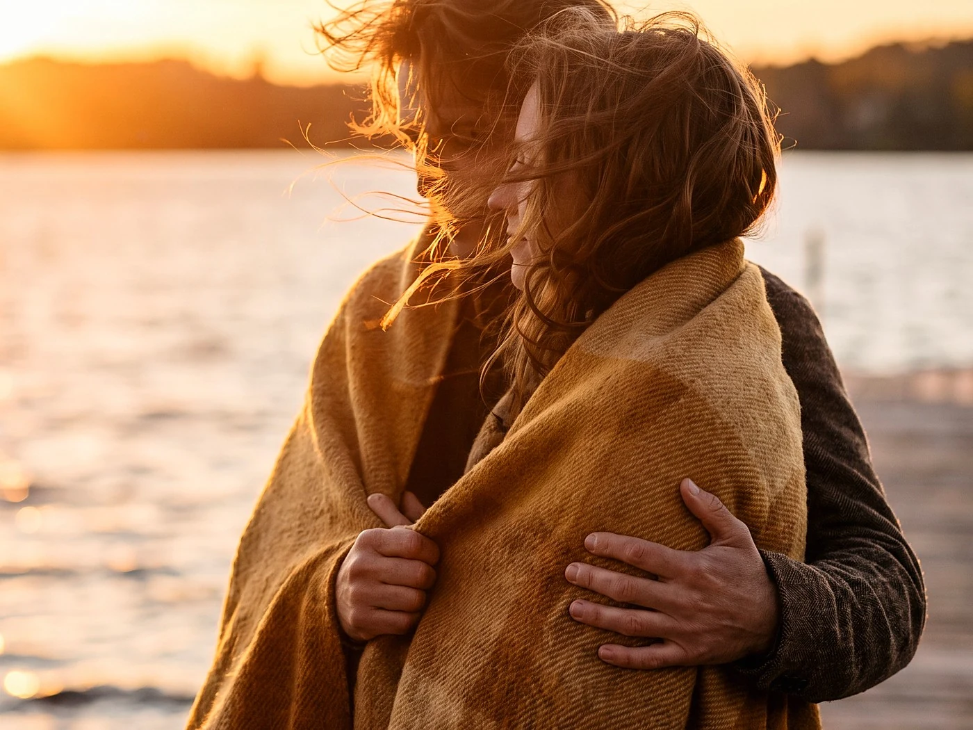 A couple wrapped in one blanket on a lakefront pier at sunset, hair moving in the wind.