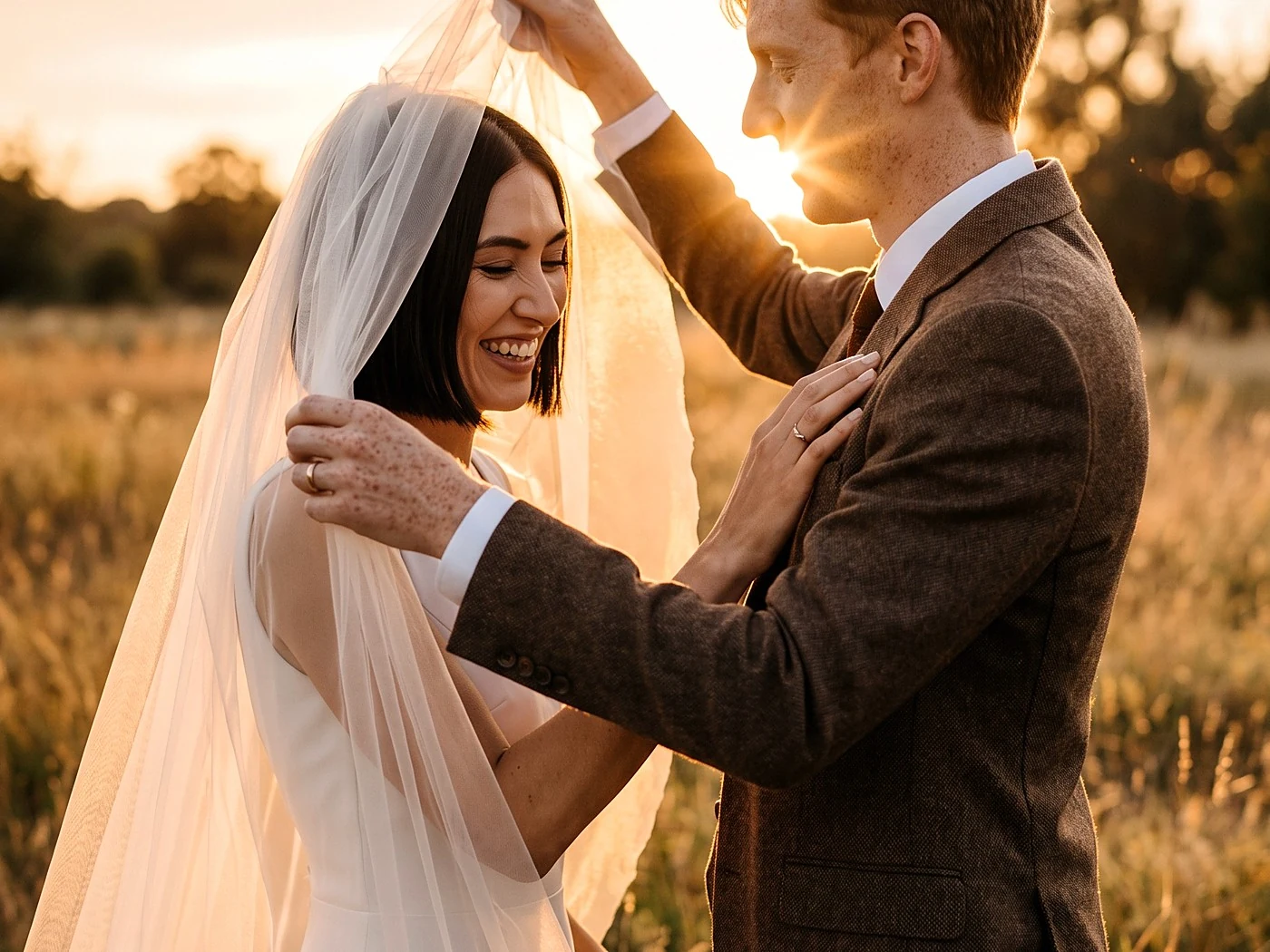 A groom in his early thirties lifting his bride's cathedral veil in a golden meadow, both laughing softly, sun.