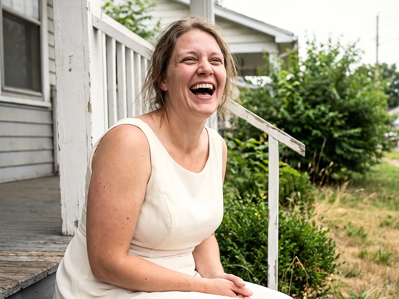 A bride laughing candidly on a porch step in a simple dress.