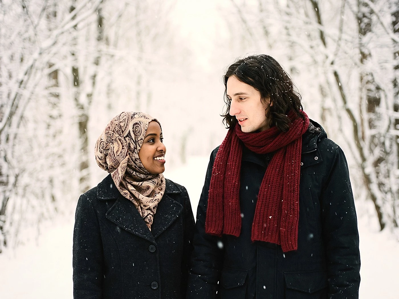 A couple walking a snowy tree tunnel, one dark coat one red scarf, flakes in the air.