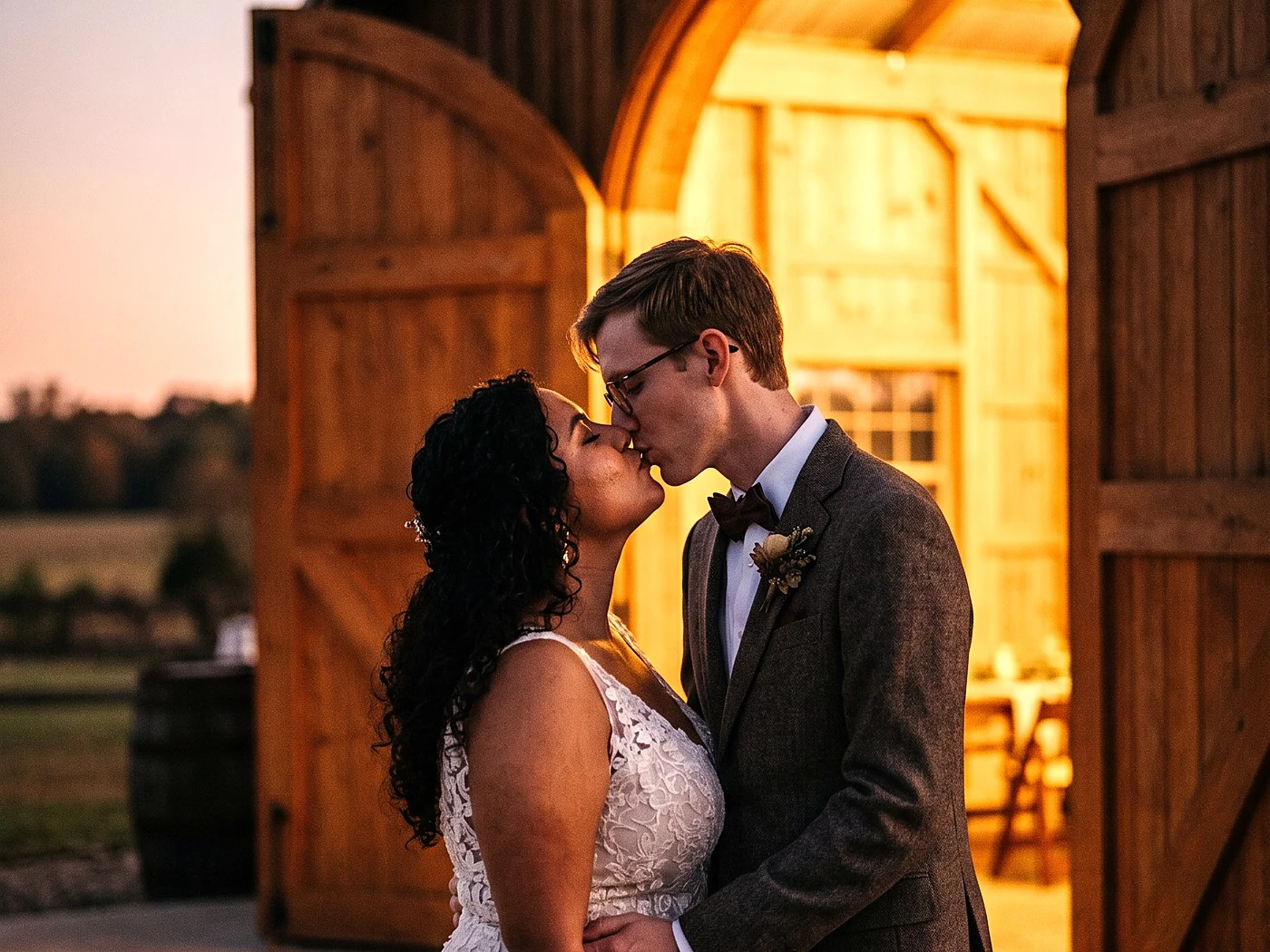A couple kissing framed by huge open barn doors, warm interior glow behind them, dusk outside.