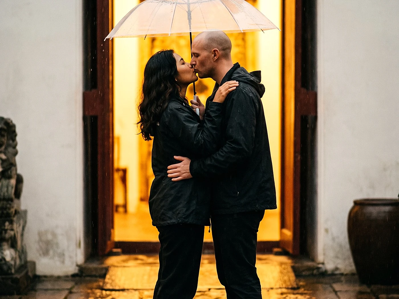 A couple kissing under a clear umbrella in light rain, wet stone path glowing golden from a doorway light.