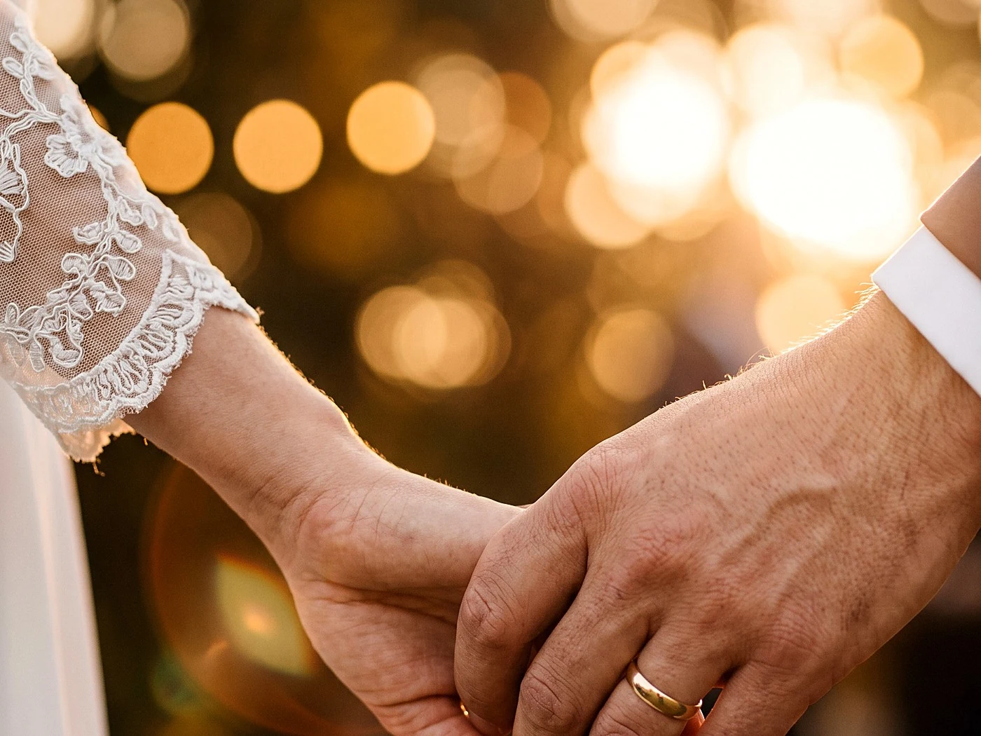 Close-up of a couple's joined hands showing both wedding bands, her sleeve lace visible, warm bokeh.